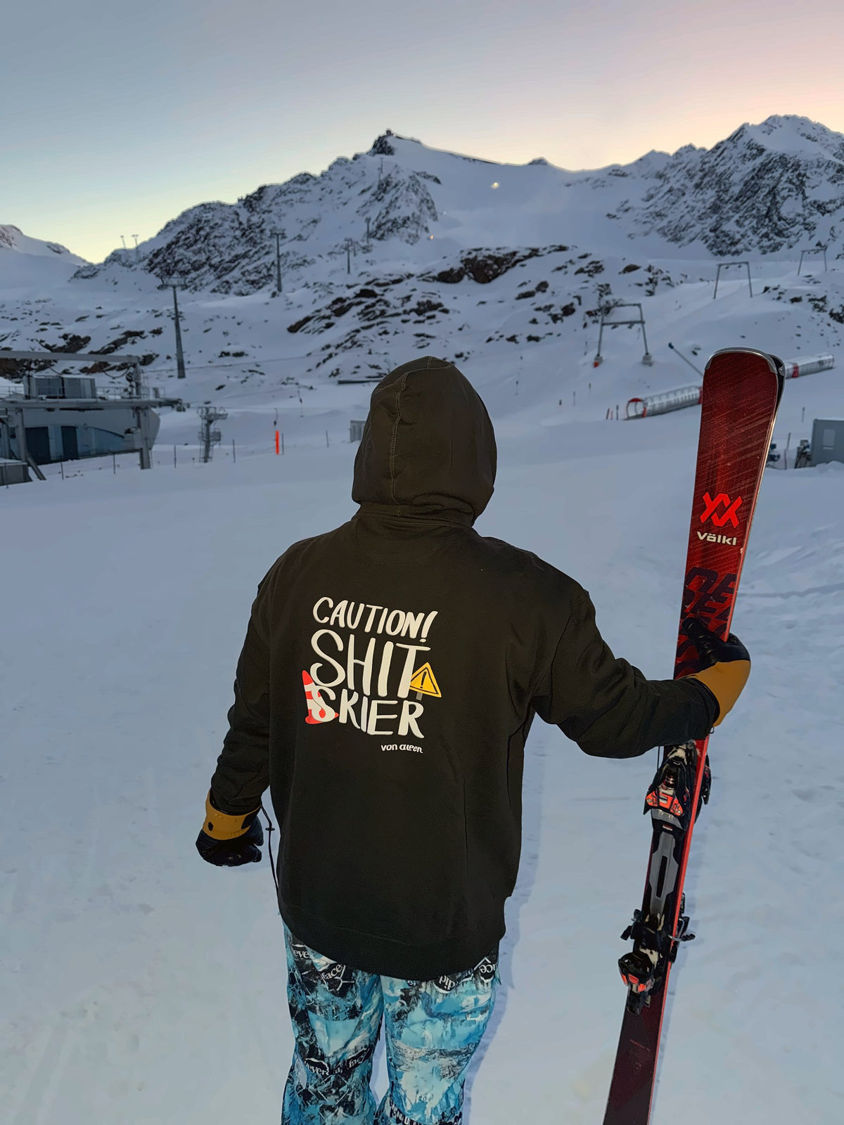 Von Alpen "Caution! Shit Skier" hoodie on the slopes at dusk, holding skis